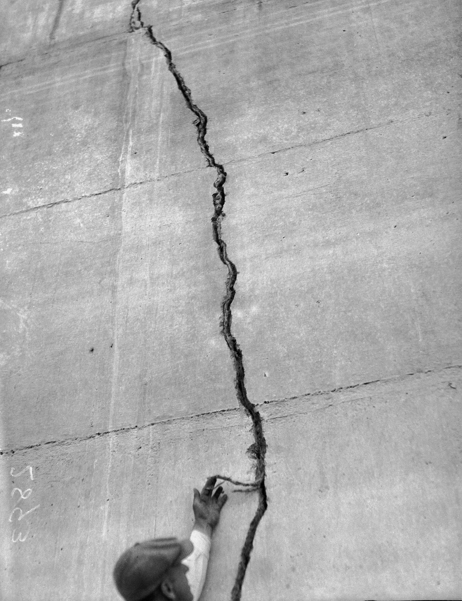 A man reaching up to a crack running down a concrete face of the St. Francis Dam, photographed after the March 1928 collapse.