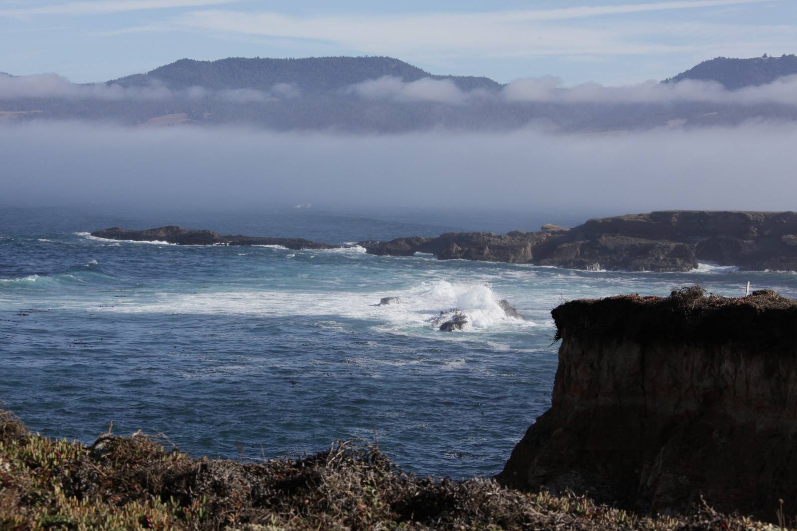 A dark bluff dropping into surf on the Stornetta Public Lands near Point Arena, low reefs breaking offshore, and a bank of fog lying along the coast beneath a ridge of mountains.