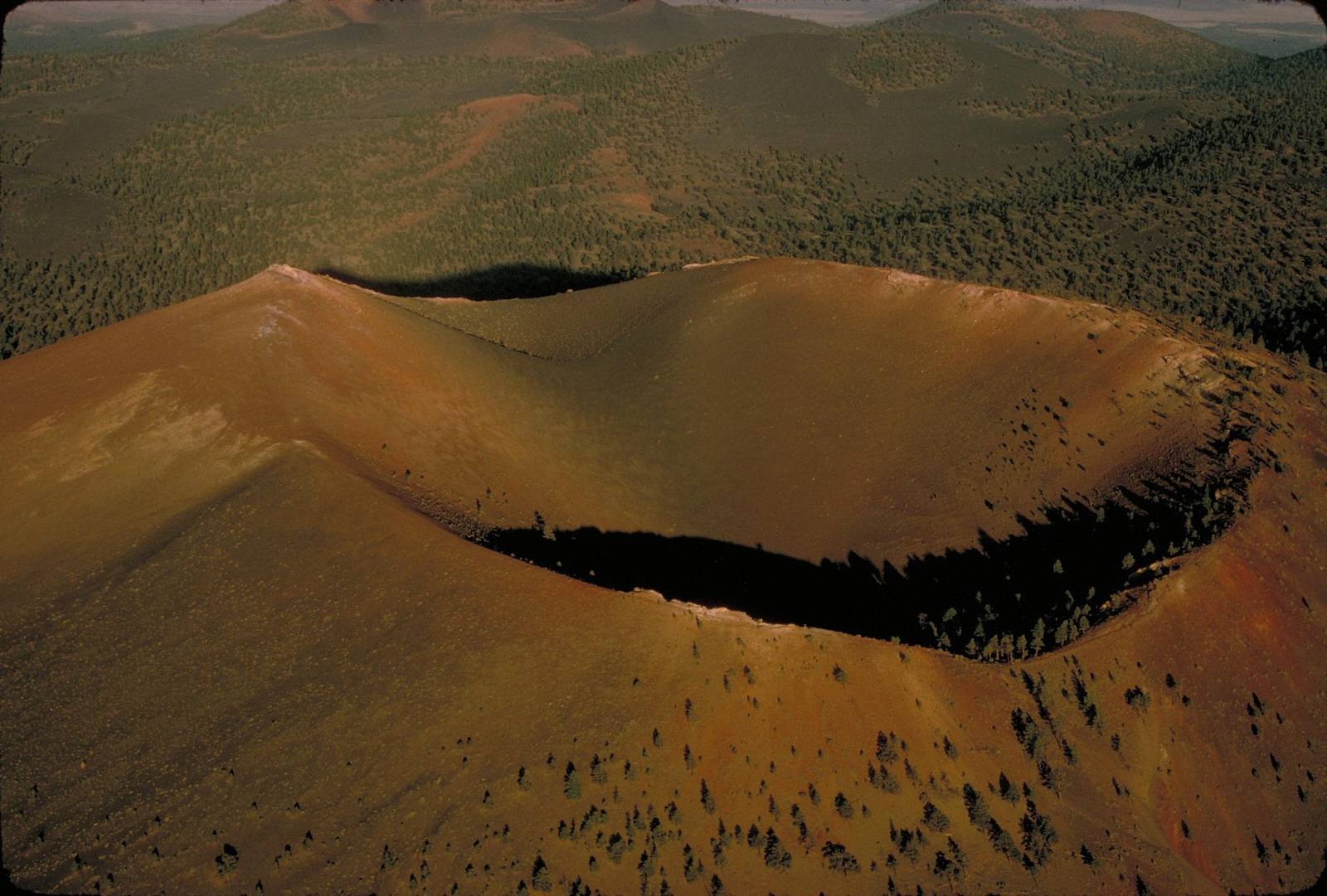 Sunset Crater's cinder cone from the air, looking down into the crater bowl — the summit rim curving across the middle of the frame with the bowl in deep shadow below it, the cone's flanks falling away in red, orange and dark brown cinder streaked with pale ash. Scattered ponderosa pines grow thinly up the slopes and stand denser along the rim. Beyond the cone the volcanic field runs back in low dark hills and green forest to a hazy horizon.