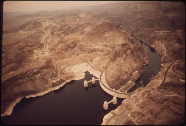 Aerial view of Hoover Dam curving across Black Canyon, Lake Mead held back upstream and the Colorado River continuing downstream through bare desert mountains.