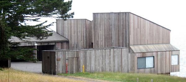 Condominium One at The Sea Ranch: a cluster of tall timber volumes clad in vertical weathered redwood boards with single-pitch roofs, set on a meadow beside cypress trees.