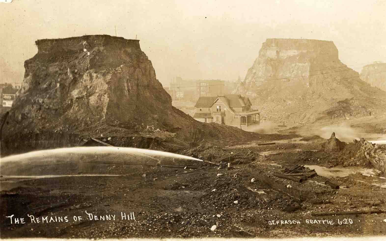 A 1920 postcard captioned The Remains of Denny Hill: two flat-topped towers of earth left standing, a house on one, while a hydraulic jet washes the ground away below