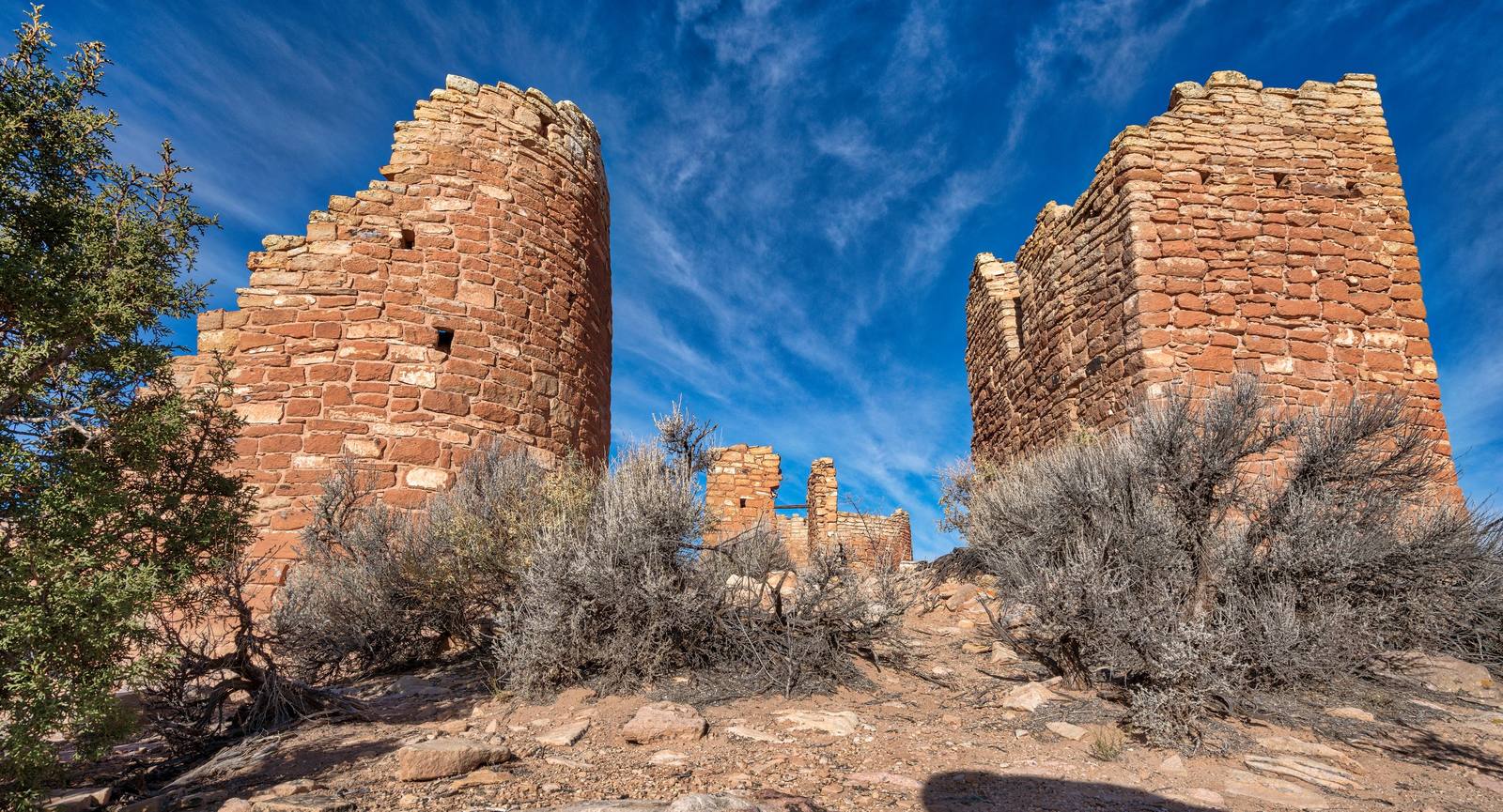 Hovenweep Castle, built by Ancestral Puebloan masons on the canyon rim in the 1200s, seen from below against a deep blue sky streaked with cirrus. Two towers stand at either side of the frame — the left one round-walled and stepping down in a broken parapet, the right one presenting a flat face and a squared corner — both laid up in coursed sandstone with small flat chinking stones packed into the joints and narrow square openings set high in the walls. A lower standing structure shows between them in the middle distance. A juniper leans in from the left, grey sagebrush covers the rocky rim across the foreground, and bare slickrock and broken red stone fill the bottom of the frame.