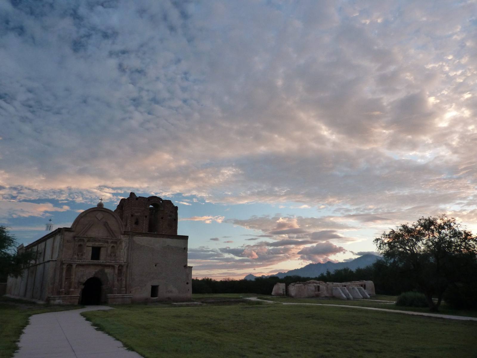The church at Tumacácori at dusk — the façade standing in shadow at the left of the frame with a curved pediment, a cross and ball finial above it, statue niches flanking an upper opening and a dark arched doorway below, and the bell tower beside it left unfinished in bare unplastered brick. A low buttressed adobe range stands in ruin to the right beyond a mesquite, a range shows low and blue on the horizon, and broken altocumulus lit pink and gold fills two thirds of the frame. A concrete path curves in across mown grass in the foreground.