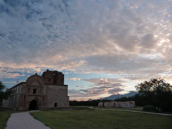 The church at Tumacácori at dusk — the façade standing in shadow at the left of the frame with a curved pediment, a cross and ball finial above it, statue niches flanking an upper opening and a dark arched doorway below, and the bell tower beside it left unfinished in bare unplastered brick. A low buttressed adobe range stands in ruin to the right beyond a mesquite, a range shows low and blue on the horizon, and broken altocumulus lit pink and gold fills two thirds of the frame. A concrete path curves in across mown grass in the foreground.