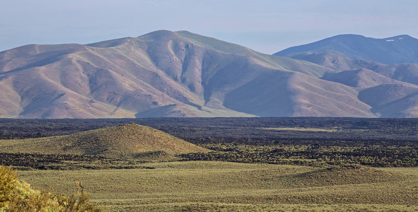 Black lava flows and a low cinder cone at Craters of the Moon, with sage-covered mountains beyond