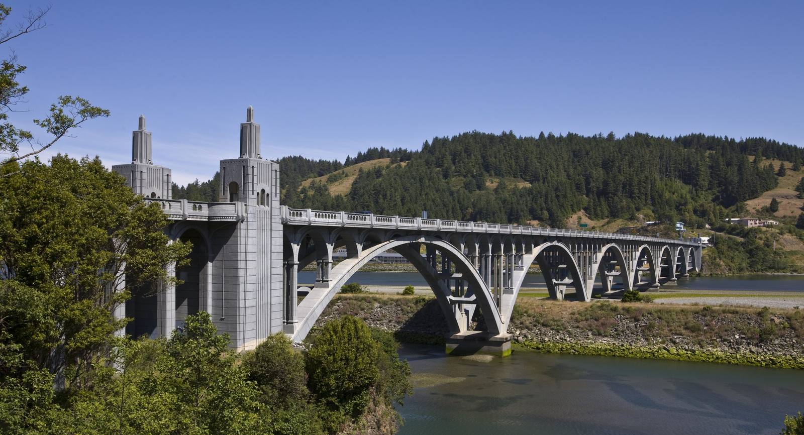 The Isaac Lee Patterson Bridge carrying US-101 over the Rogue River at Gold Beach, seen from the southeast: a pair of tall Art Deco concrete pylons at the near end, then a run of open-spandrel arches stepping across the river to a forested hillside with a few houses on the slope. Trees frame the left edge, the river's green water fills the foreground, and the sky is a clear deep blue.
