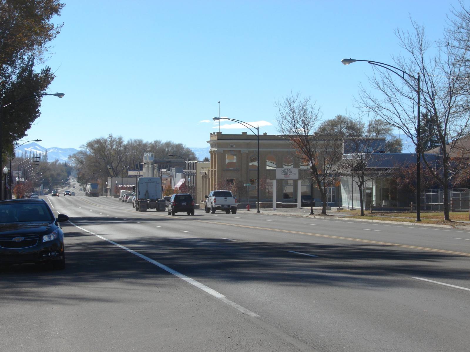 US-89 running as Gunnison's main street, four wide lanes receding toward snow-dusted mountains on the horizon. A dark sedan sits at the near left edge, with a white pickup, a dark SUV and a box truck further down the road. At right stands a two-storey commercial block of tan stone with a shaped parapet and tall arched upper windows, a hanging sign in front of it lettered DAVES CUSTOM CABINETS, and low white storefronts beyond. Bare trees and curved street lamps line both kerbs under a pale blue sky.