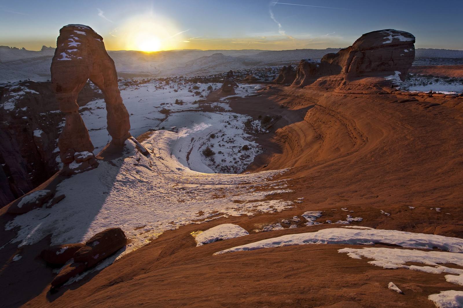 Delicate Arch in Arches National Park under snow, standing on the rim of its slickrock bowl with the low sun on the horizon just to the right of the opening. Snow lies along the arch's shaded ledges and across the bowl below, a snow-streaked sandstone fin rises at right, and distant mesas break the skyline. Of the five Utah national parks this itinerary covers, only Arches is in the frame.
