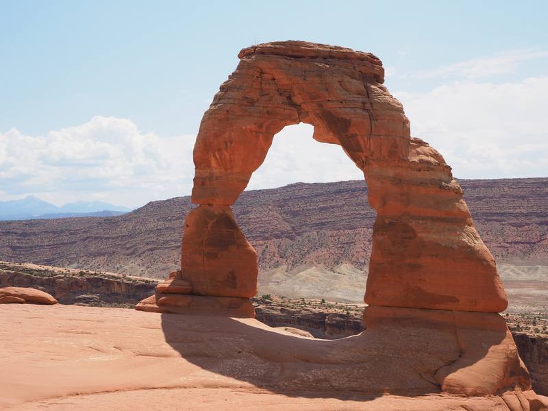 Delicate Arch in Arches National Park, standing free on the rim of its slickrock bowl — a single span of red sandstone with one thick leg and one slender, streaked with dark desert varnish and banded pale cream along its upper curve, throwing a hard shadow across the smooth rock at its foot. Behind it the country falls away in red and grey ledges to a broad bench, and the La Sal Mountains stand hazy blue on the horizon at the left under scattered cloud.