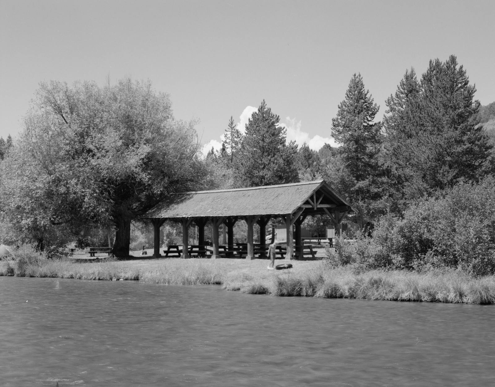 The Warm River sliding past a timber picnic shelter and willows at Warm River Campground, a tuber standing on the grassy bank, in a Historic American Buildings Survey photograph