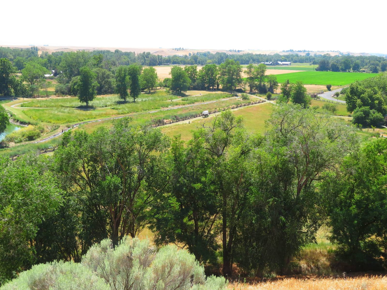 The grounds of Whitman Mission National Historic Site seen from the hill above, cottonwoods along a water channel and green farm fields beyond